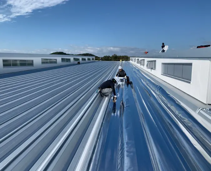 青空のもと、建物の折板屋根上で施工作業を行う作業員たち